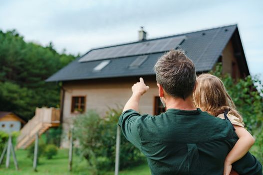 hombre sosteniendo a su hija mientras señala las placas solares de su tejado